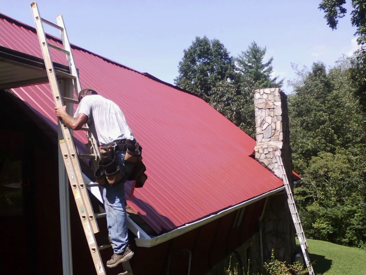 Roofer on a ladder beside a red metal roof and stone chimney — Soffit & Fascia Repair by Roofing Brothers in Loa